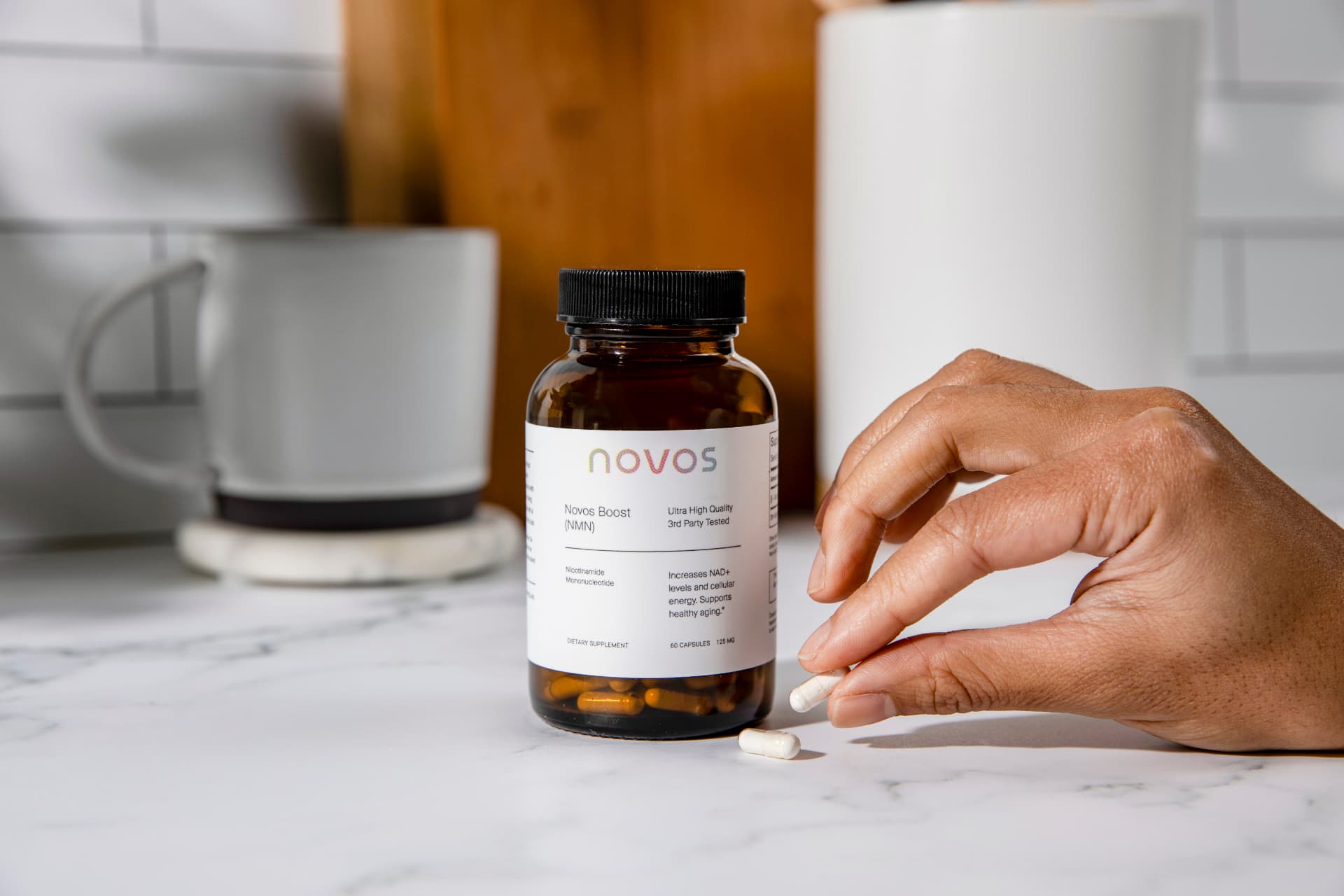 Hand reaching for a NOVOS Boost NMN capsule beside the bottle on a kitchen counter