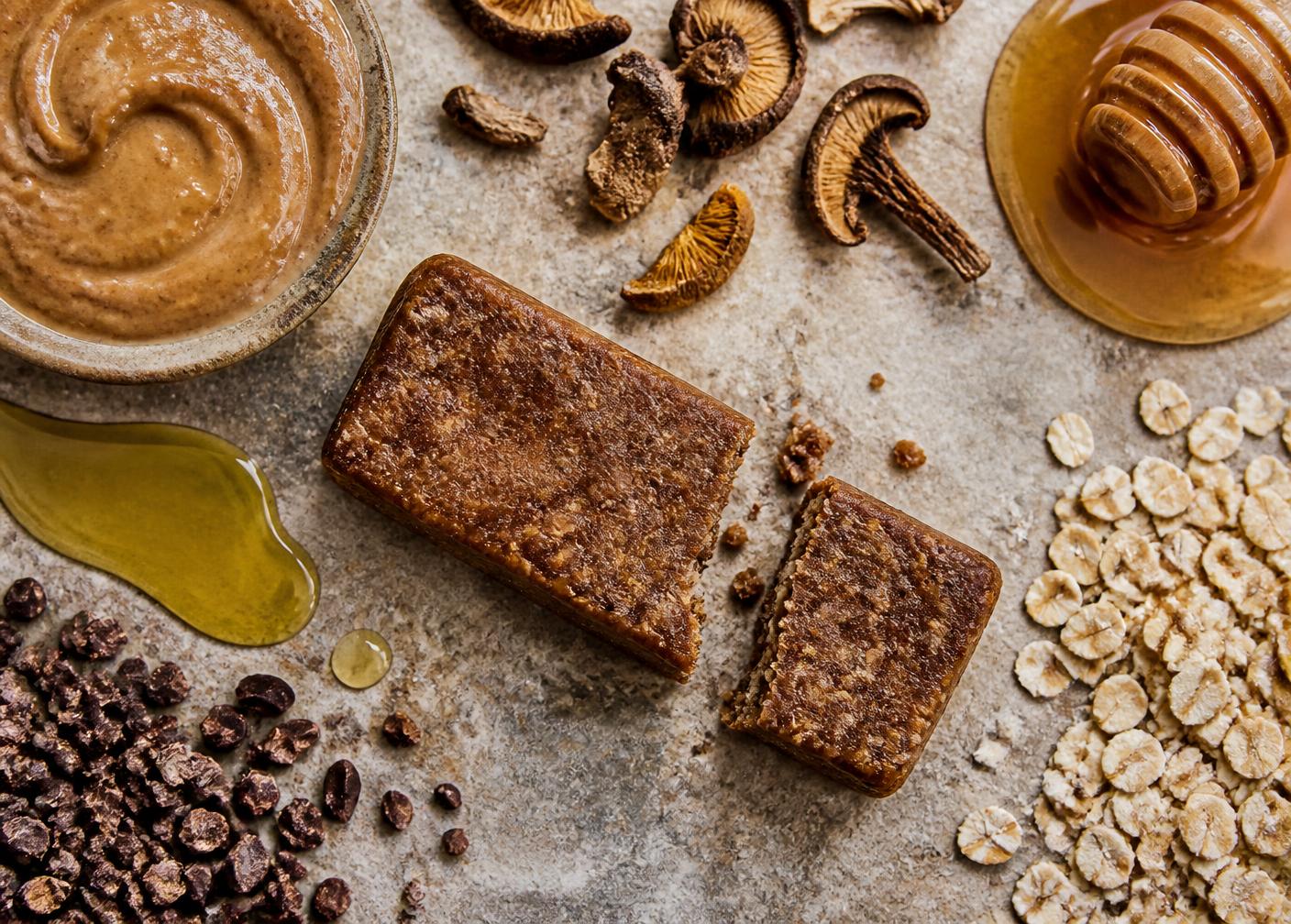 A NOVOS Bar broken open beside seed butter, olive oil, cocoa, mushrooms, honey and oats